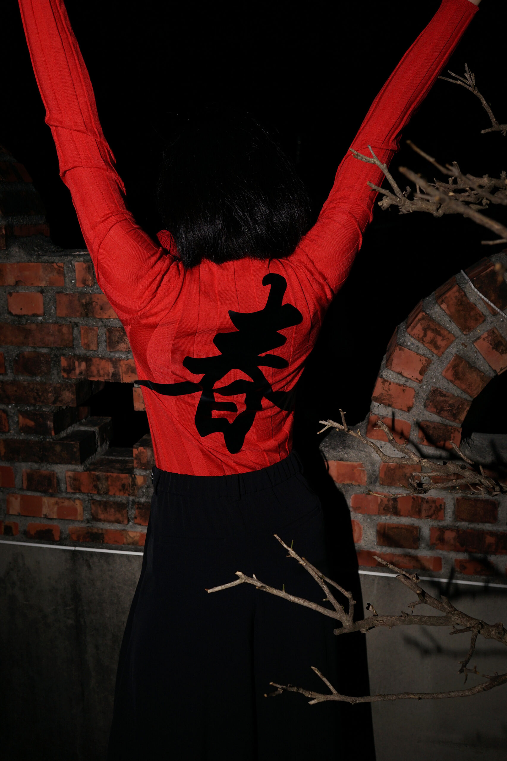 Girl wears Red jacket with asian character on the back with both arm raised, new a dark brick background
